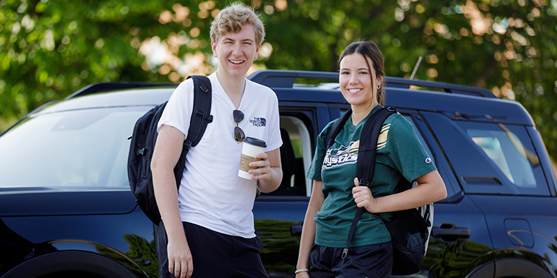Two college age kids standing in front of a vehicle with coffee in their hands
