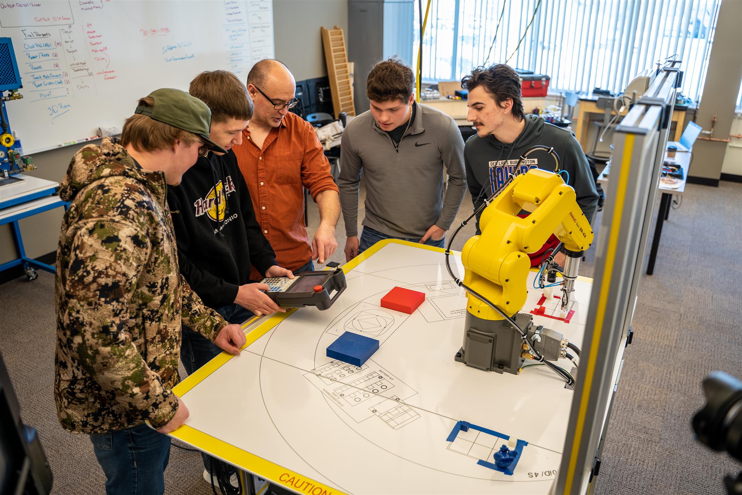Group of mechatronic students and an instructor standing around a drafting table and yellow automation arm.